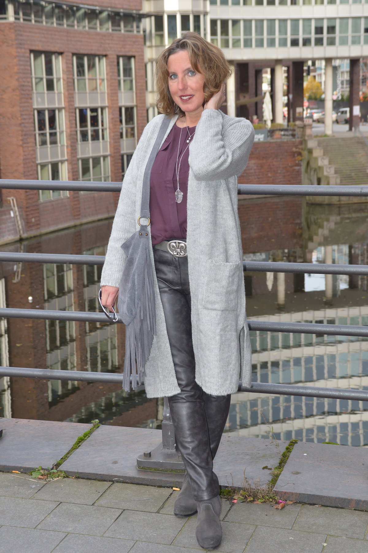 Ari Sunshine kombiniert einen langen grauen Cardian zur pflaumefarbenen Bluse, schicker Lederimitat-Hose und grauen Boots.