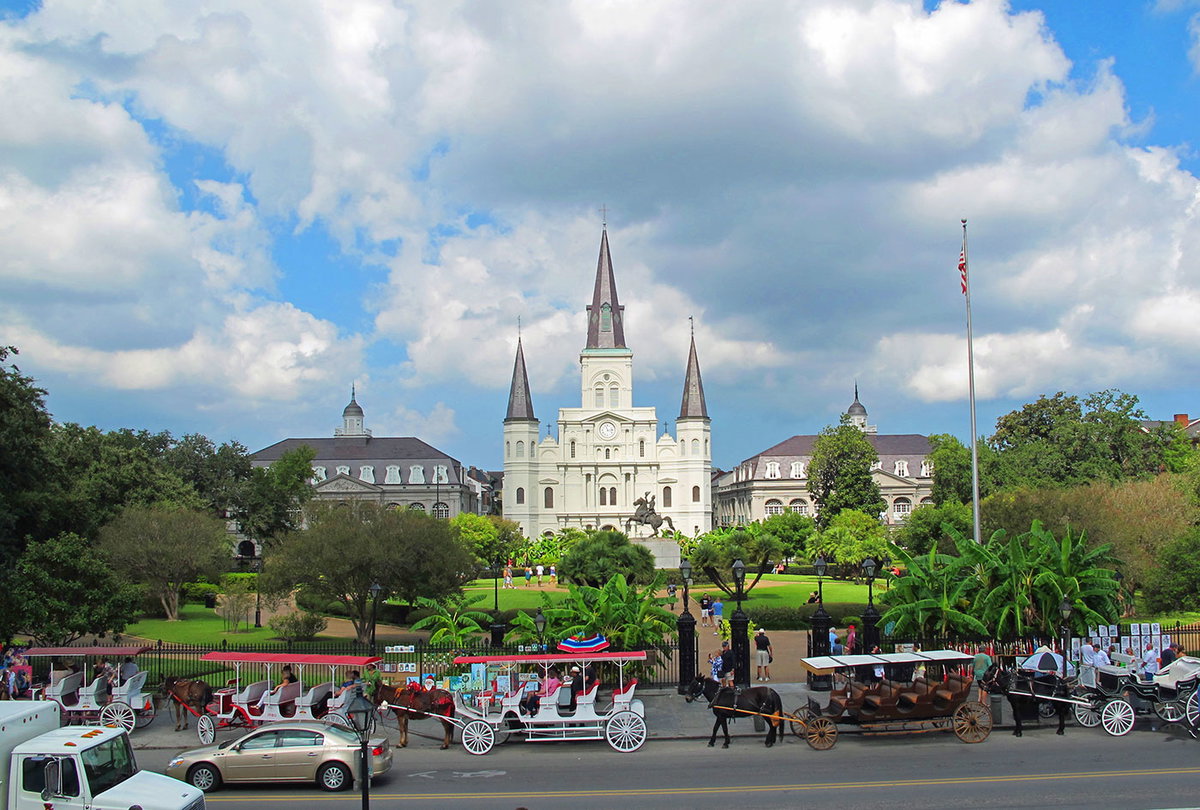 Begrünter Platz namens "Jackson Square" mit rot-weißen Pferdekutschen davor und der weißen St.-Louis-Kathedrale mit drei spitzen Türmen