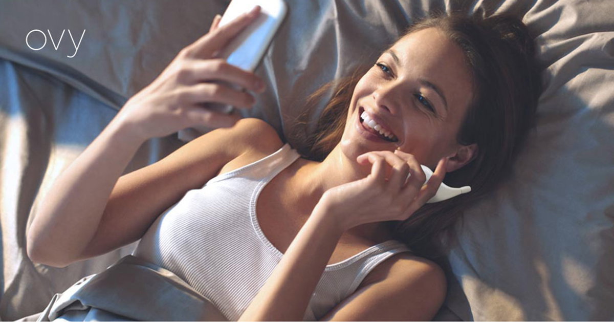 Frau liegt im Bett und blickt glücklich auf ihr Smartphone, in der anderen Hand hält sie ein Thermometer