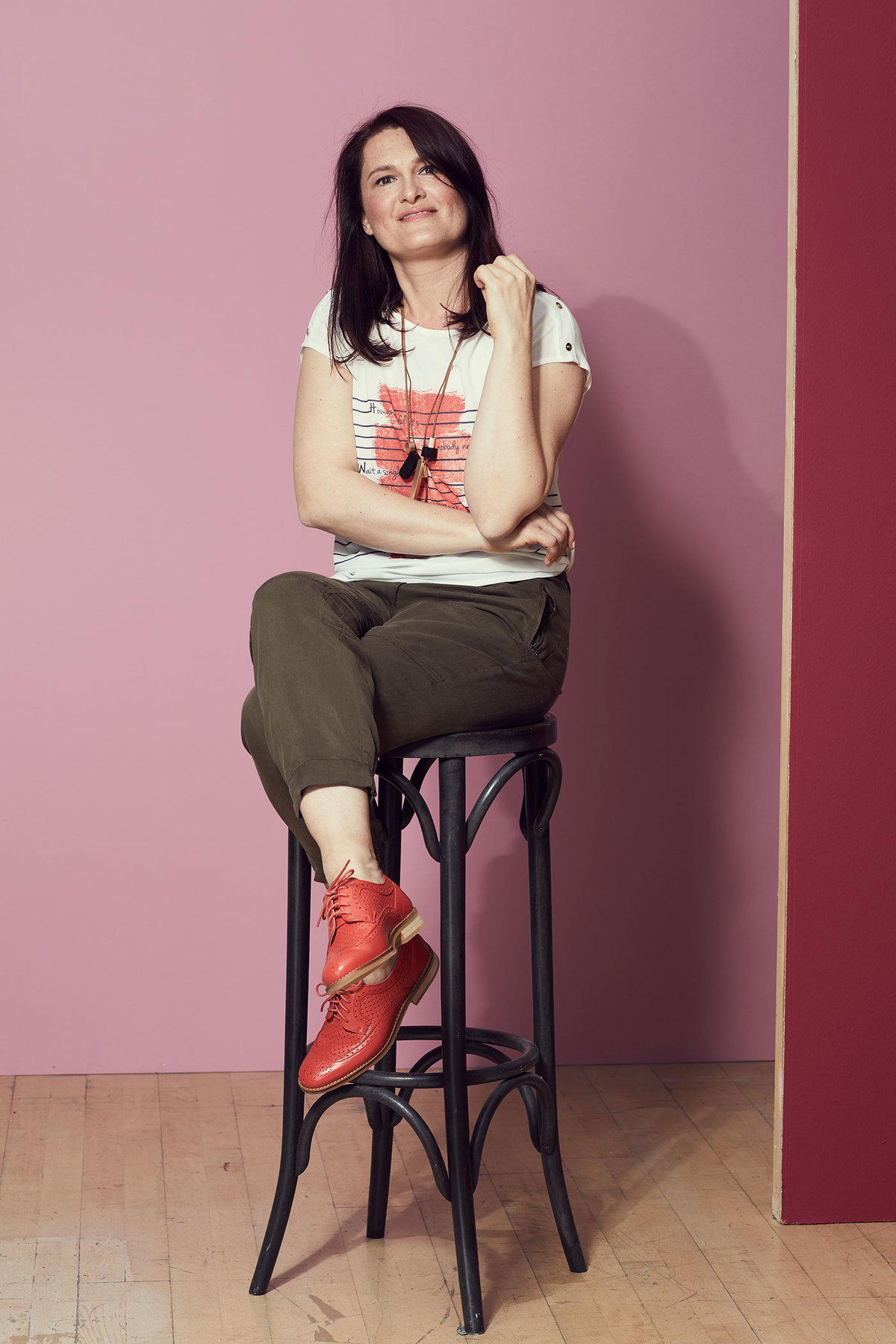Inga sitzt auf einem Barhocker mit khakifarbener Hose und korallroten Schuhen passend zum T-Shirt mit Aufdruck