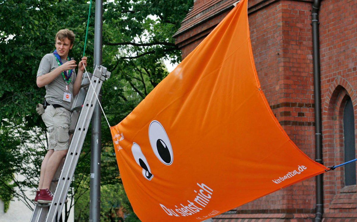 Junge in Pfadfinderuniform steht auf Leiter und befestigt ein orangefarbenes Banner