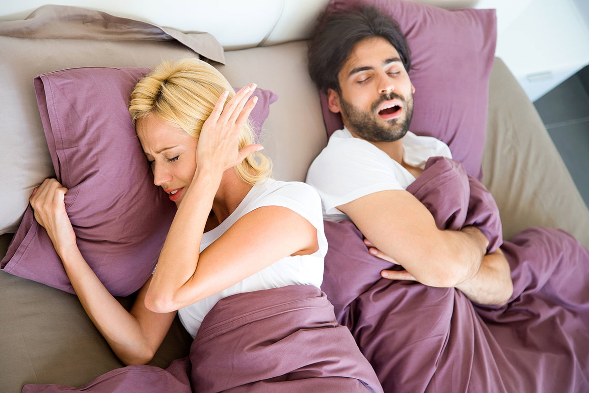 Frau liegt neben schlafendem Mann im Bett und hält sich ein Ohr zu.