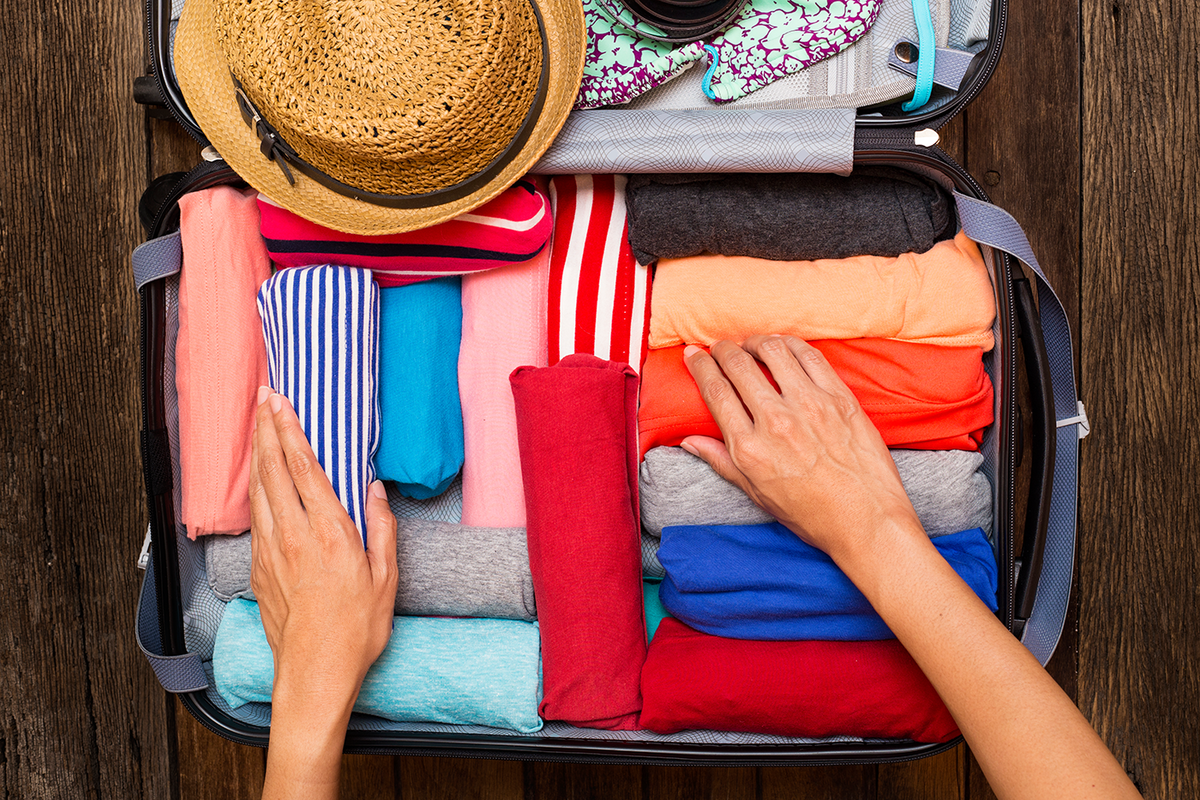 Koffer mit gerollten, bunten T-Shirts, einem Strohhut und einem Bikini